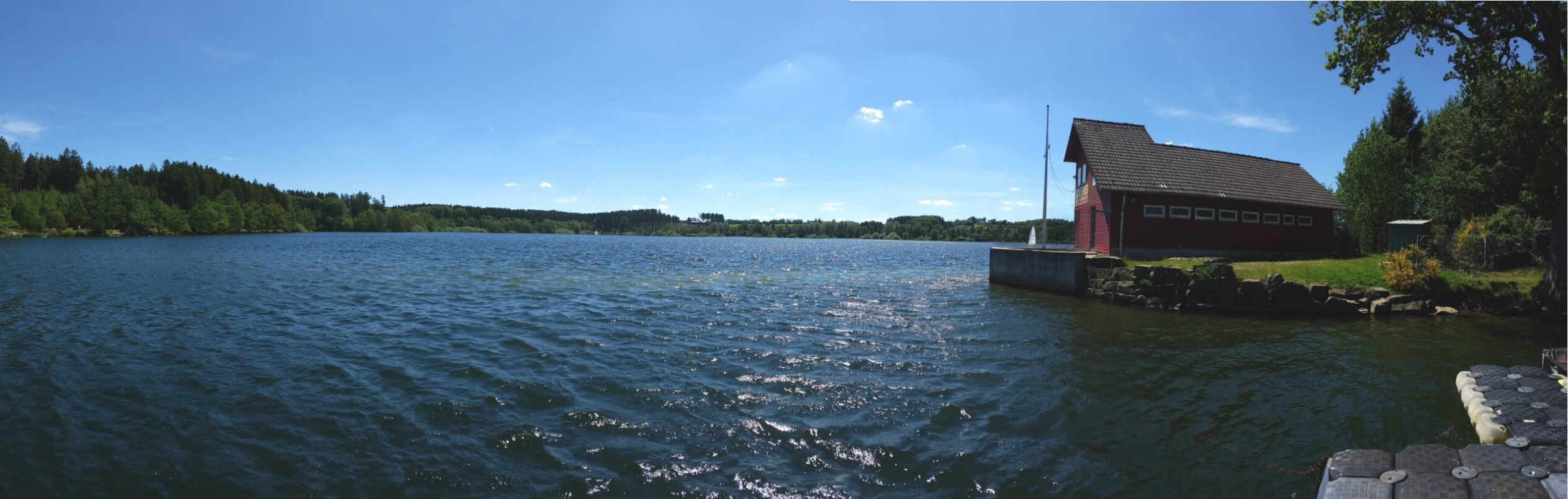 Die Brucher Talsperre mit bewaldetem Ufer und Bootshaus bei blauem Himmel